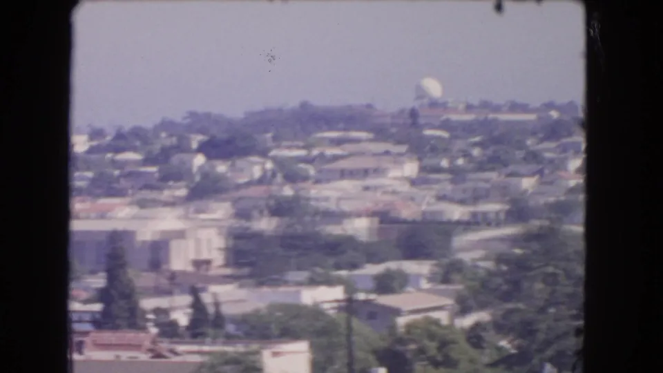 1966 Los Angeles 8mm frame showing a hazy residential hillside with clustered houses, trees, rooftops, and a distant white dome-like structure.
