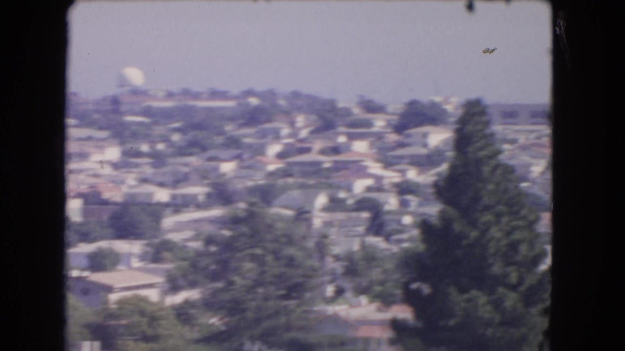 1966 Los Angeles home-movie frame looking over residential rooftops and trees toward a distant white dome and hazy hillside.