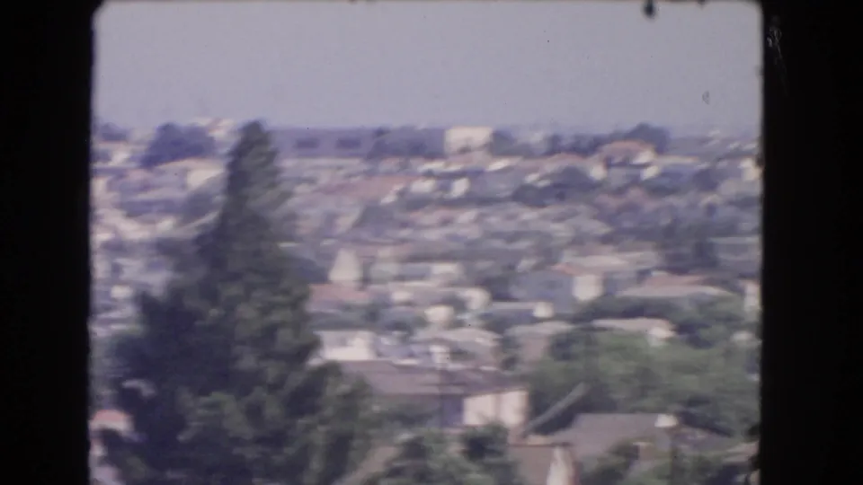 1966 Los Angeles 8mm frame showing a soft, drifting aerial view of hillside homes, trees, rooftops, and a prominent conifer.