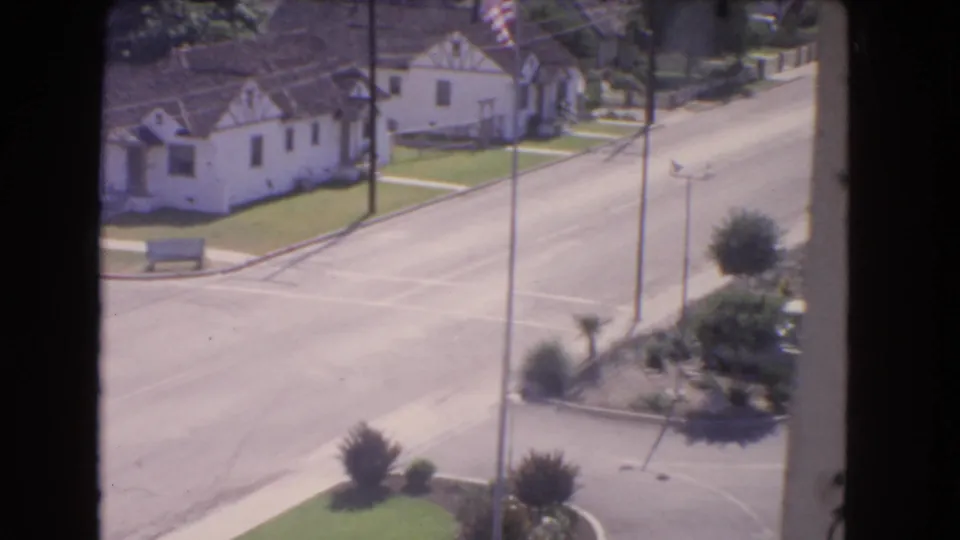 1966 Los Angeles residential street view with mid-century houses, lawns, utility poles, an intersection, and a small American flag.