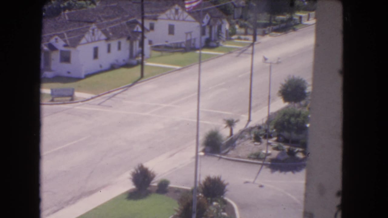 Elevated view of a quiet street corner with white stucco houses, lawns, utility poles, and shrubs.