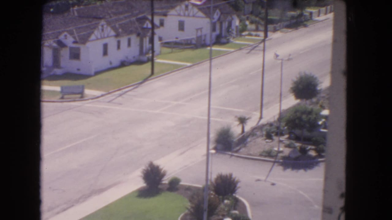 1966 Los Angeles view over a residential street, showing stucco homes, tile roofs, lawns, utility poles, and a planted corner near the roadway.