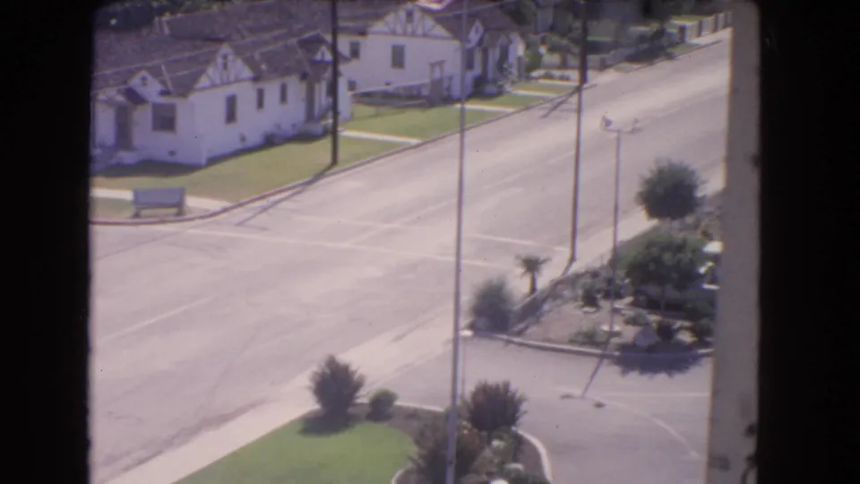 1966 Los Angeles view over a residential street, showing stucco homes, tile roofs, lawns, utility poles, and a planted corner near the roadway.