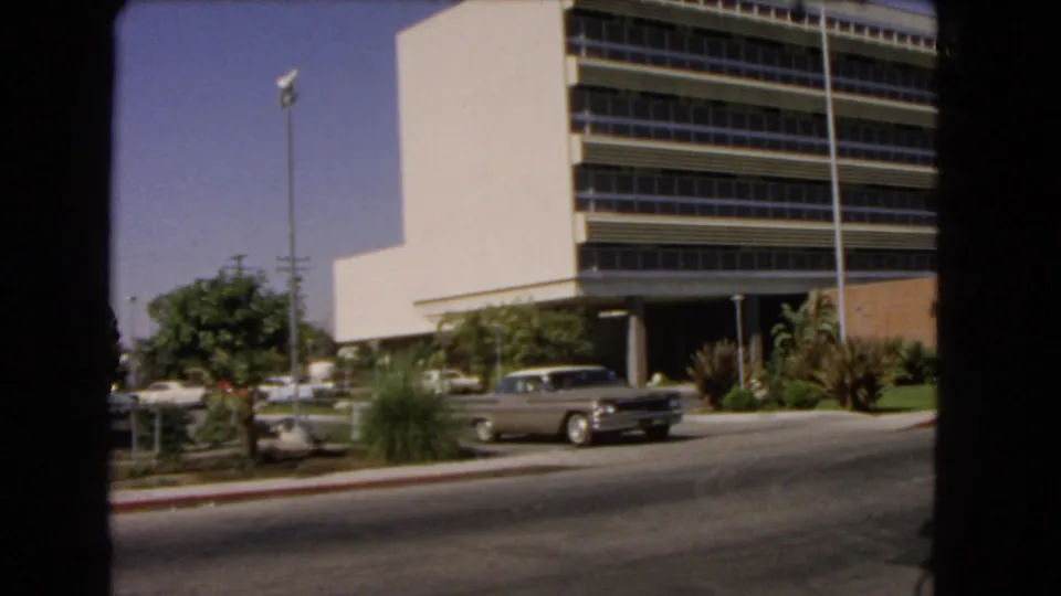 1966 Los Angeles street-level frame of a modern multi-story building with horizontal window bands, landscaping, a light pole, and parked classic cars.