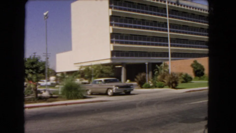 1966 Los Angeles frame showing a modern building facade with long window bands, curbside landscaping, a tall light pole, and classic cars nearby.