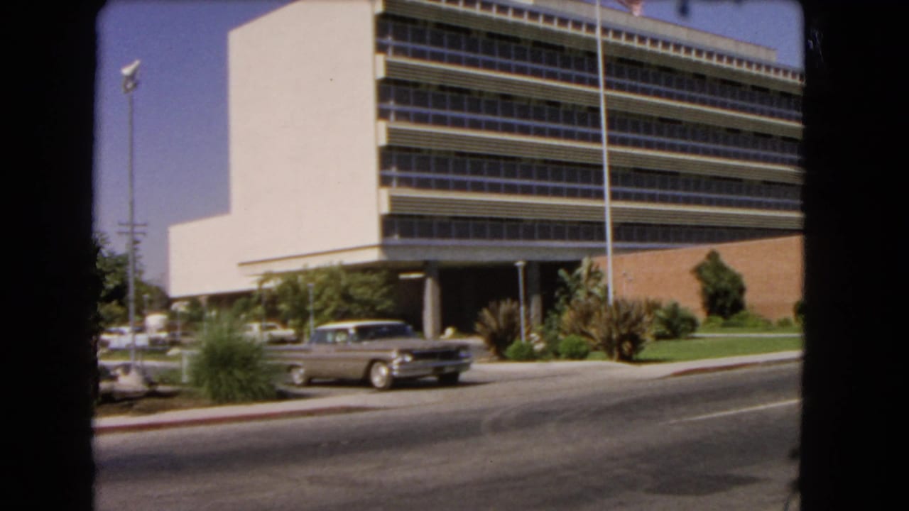 1966 Los Angeles street view with a silver sedan passing a modernist office building, flagpole, lawn, and palm landscaping.