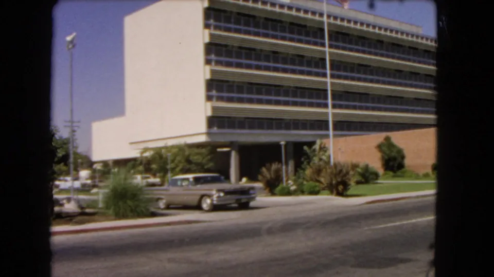 1966 Los Angeles street view with a silver sedan passing a modernist office building, flagpole, lawn, and palm landscaping.
