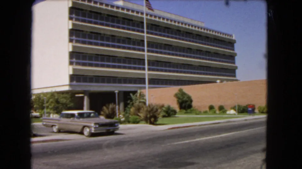 1966 Los Angeles frame with the sedan partly lower left, a modernist office block behind it, and a tall flagpole in front.