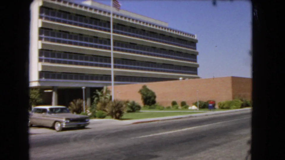 1966 Los Angeles street scene with the sedan leaving the frame and the office building, lawn, flagpole, and shrubs still visible.