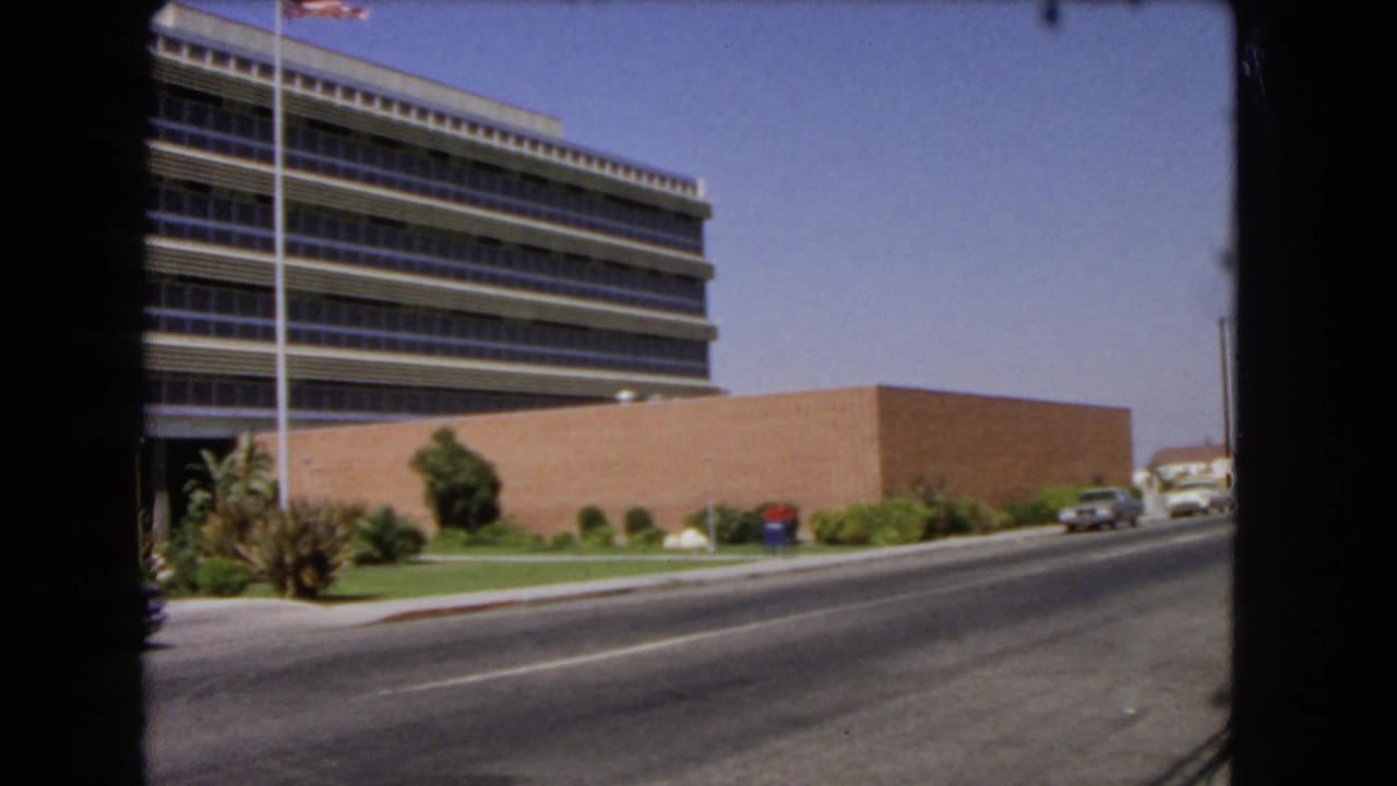 1966 Los Angeles frame with a mid-century office building, flagpole, palms, and a light-colored sedan moving along the road.