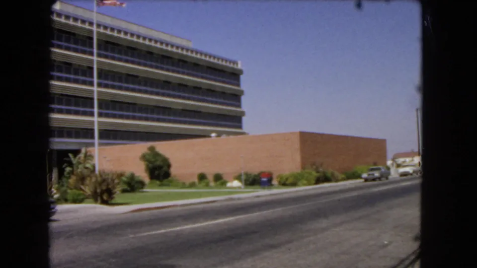 1966 Los Angeles frame with a mid-century office building, flagpole, palms, and a light-colored sedan moving along the road.
