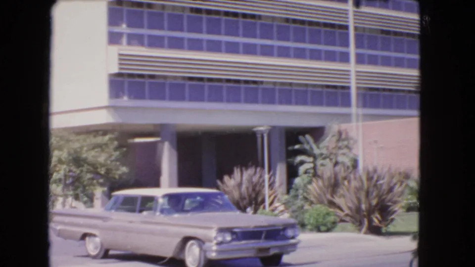 Closer 1966 Los Angeles home-movie view of the building facade, entrance columns, roadside planting, and sedan in motion.