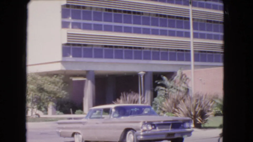 1966 Los Angeles frame showing the sedan near the right side of the road with the office building facade and planting behind it.