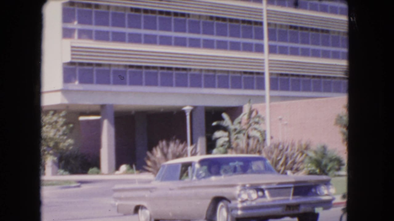 1966 Los Angeles home-movie frame showing a light-colored sedan near a modern building with purple-tinted windows and palm landscaping.