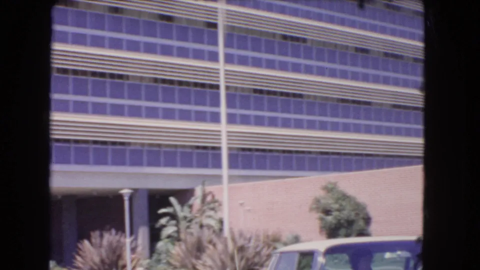 1966 Los Angeles frame with a modernist facade, purple-tinted windows, shrubs, and the edge of a car at lower right.