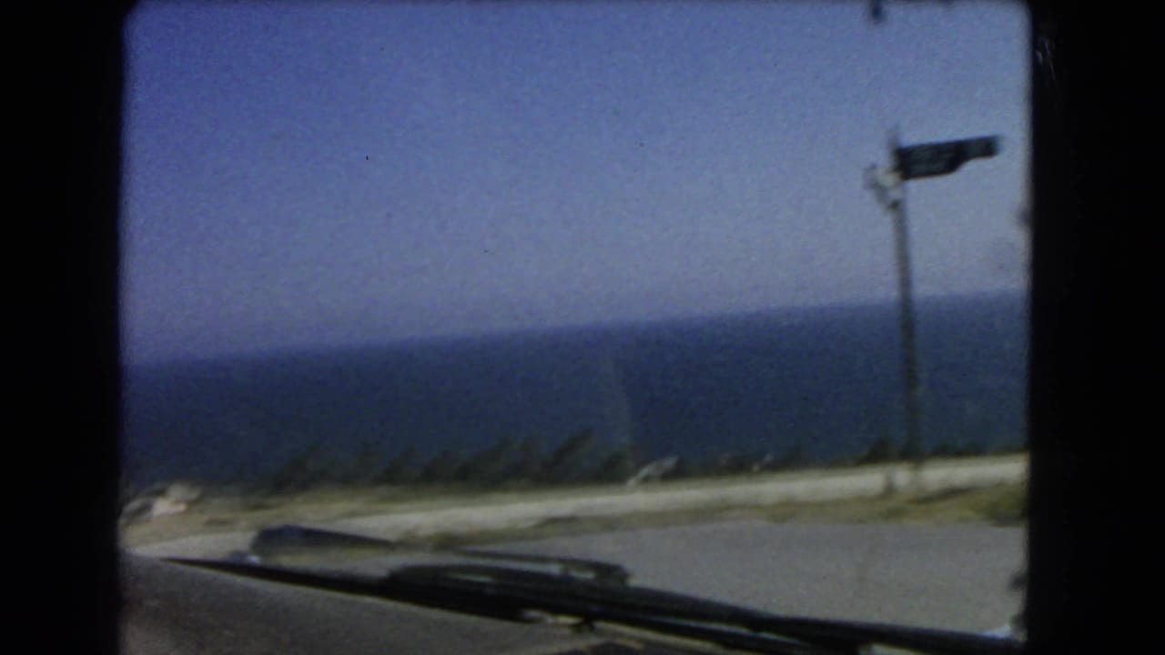 1966 Los Angeles coastal driving frame showing a blurred ocean horizon, roadside pole, and the car hood at the bottom edge.