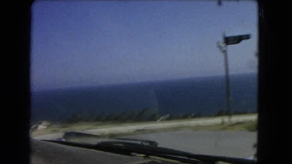 1966 Los Angeles coastal driving frame showing a blurred ocean horizon, roadside pole, and the car hood at the bottom edge.