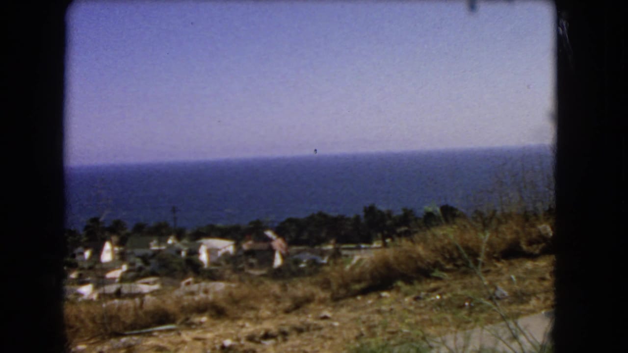 1966 Los Angeles coastal frame showing ocean, hillside vegetation, scattered buildings, and a road edge from a moving viewpoint.