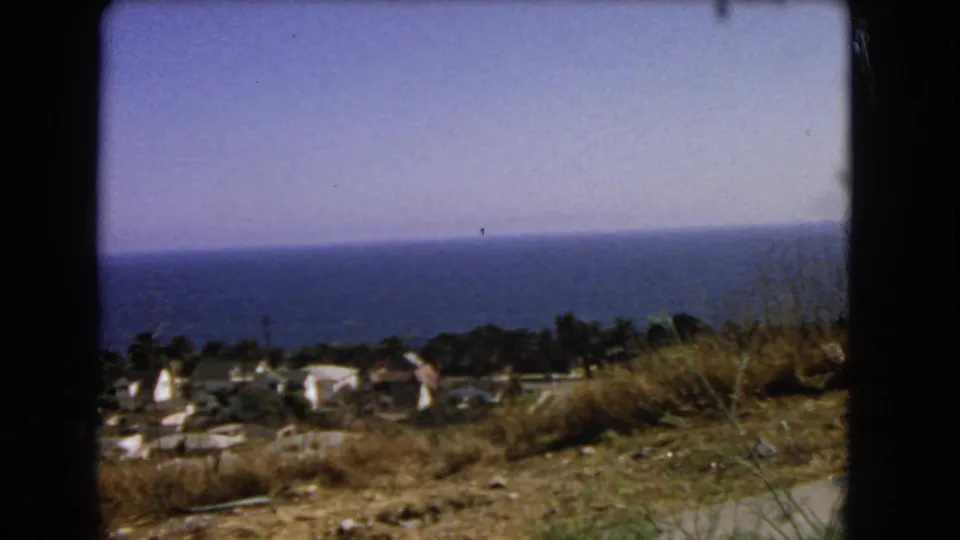 1966 Los Angeles coastal frame showing ocean, hillside vegetation, scattered buildings, and a road edge from a moving viewpoint.