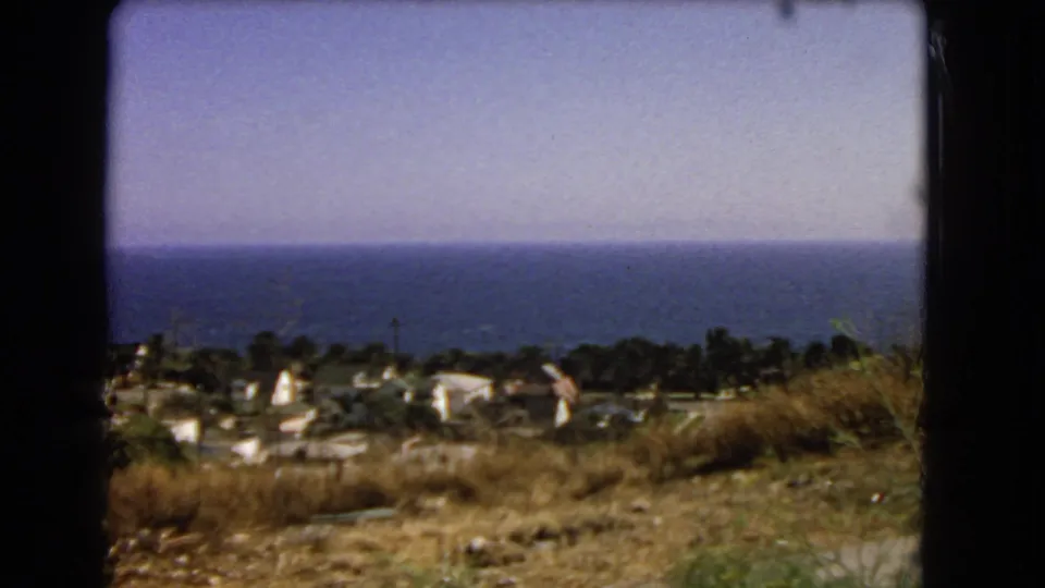 Soft 1966 8mm frame from a higher Los Angeles-area viewpoint, looking past dry ground and rooftops toward the Pacific.