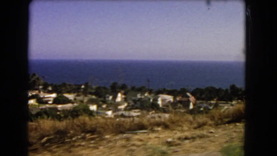 1966 Los Angeles-area home-movie frame showing dry hillside brush, small coastal buildings, trees, and the ocean under a pale sky.