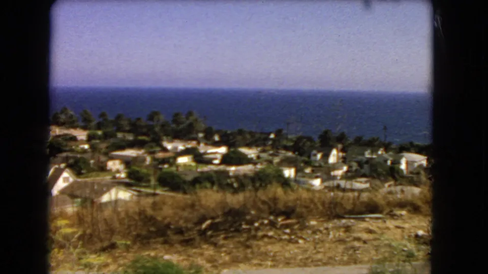 1966 Southern California coastal hillside with dry brush, light-roofed houses, palms, and the Pacific Ocean in the distance.
