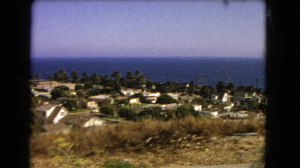 Los Angeles coastal neighborhood seen from a hillside, with dry scrub, palms, modest homes, and blue ocean in a 1966 8mm frame.
