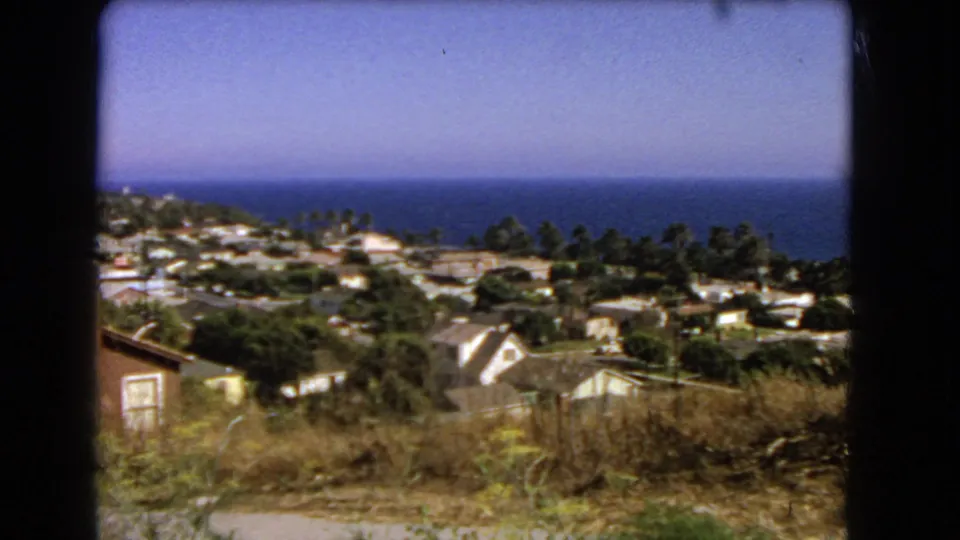 1966 Los Angeles-area frame showing a dry slope, small structure, clustered homes, trees, and the Pacific Ocean beyond.