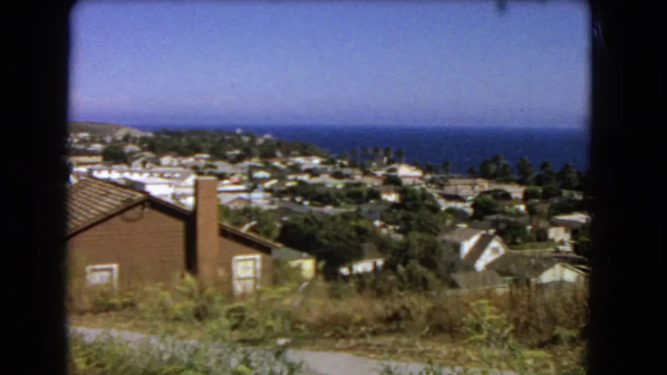 1966 Los Angeles hillside view with a brick house, chimney, dry plants, nearby roofs, trees, and blue Pacific water behind the neighborhood.