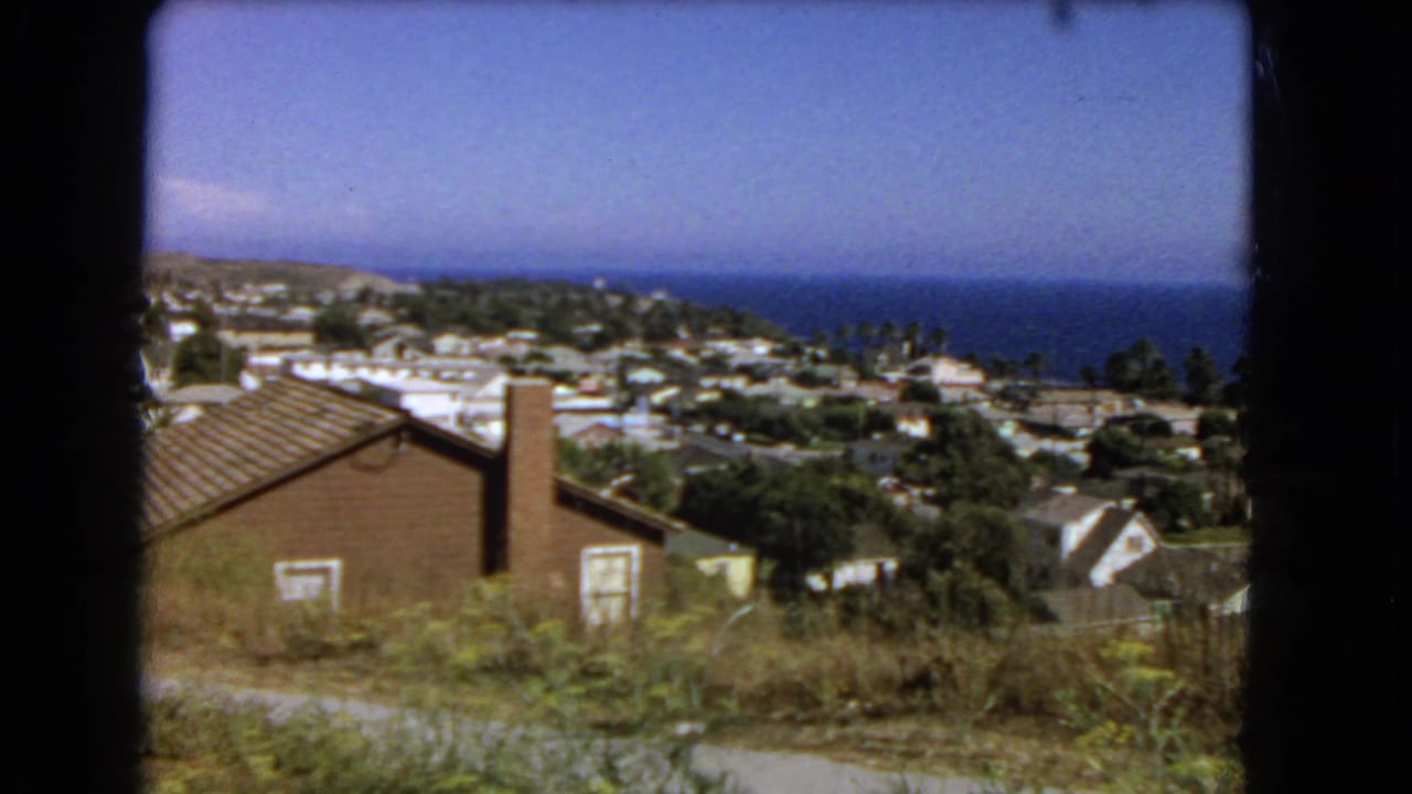 1966 Los Angeles coastal hillside view with residential rooftops, foreground brush, and the Pacific horizon in the distance.