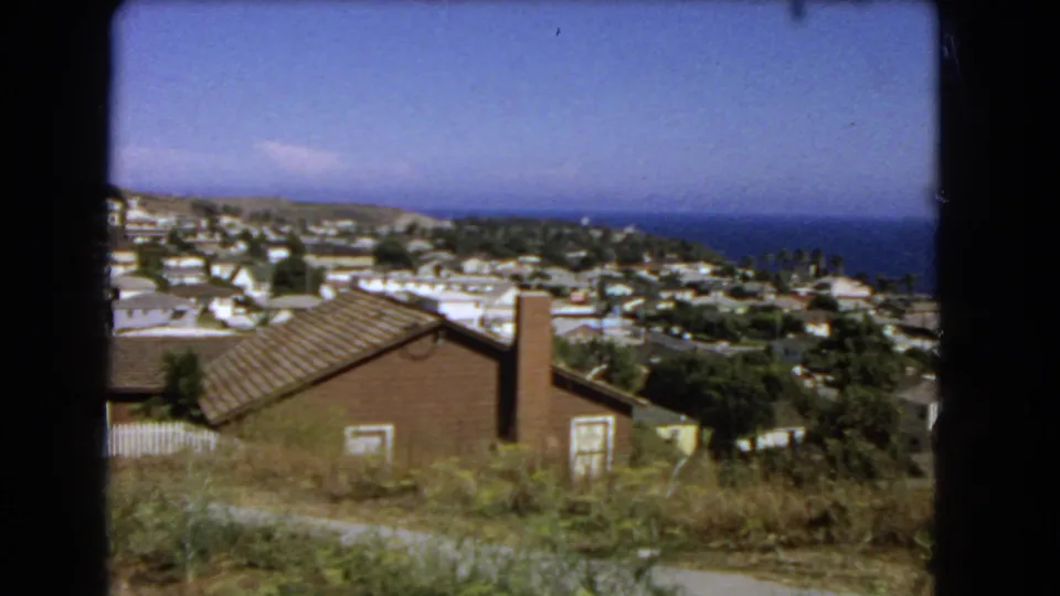 8mm frame of a coastal Los Angeles hillside neighborhood, showing rooftops, foreground vegetation, roads, and ocean horizon.