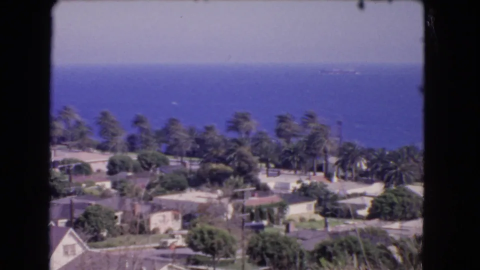 1966 home-movie frame of Los Angeles coastal homes and palms below a hillside viewpoint, with ocean beyond.