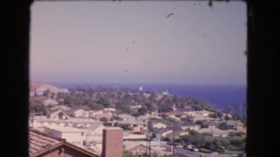 1966 coastal Los Angeles view with a foreground roof and chimney, dense homes, trees, and the distant Pacific coastline.