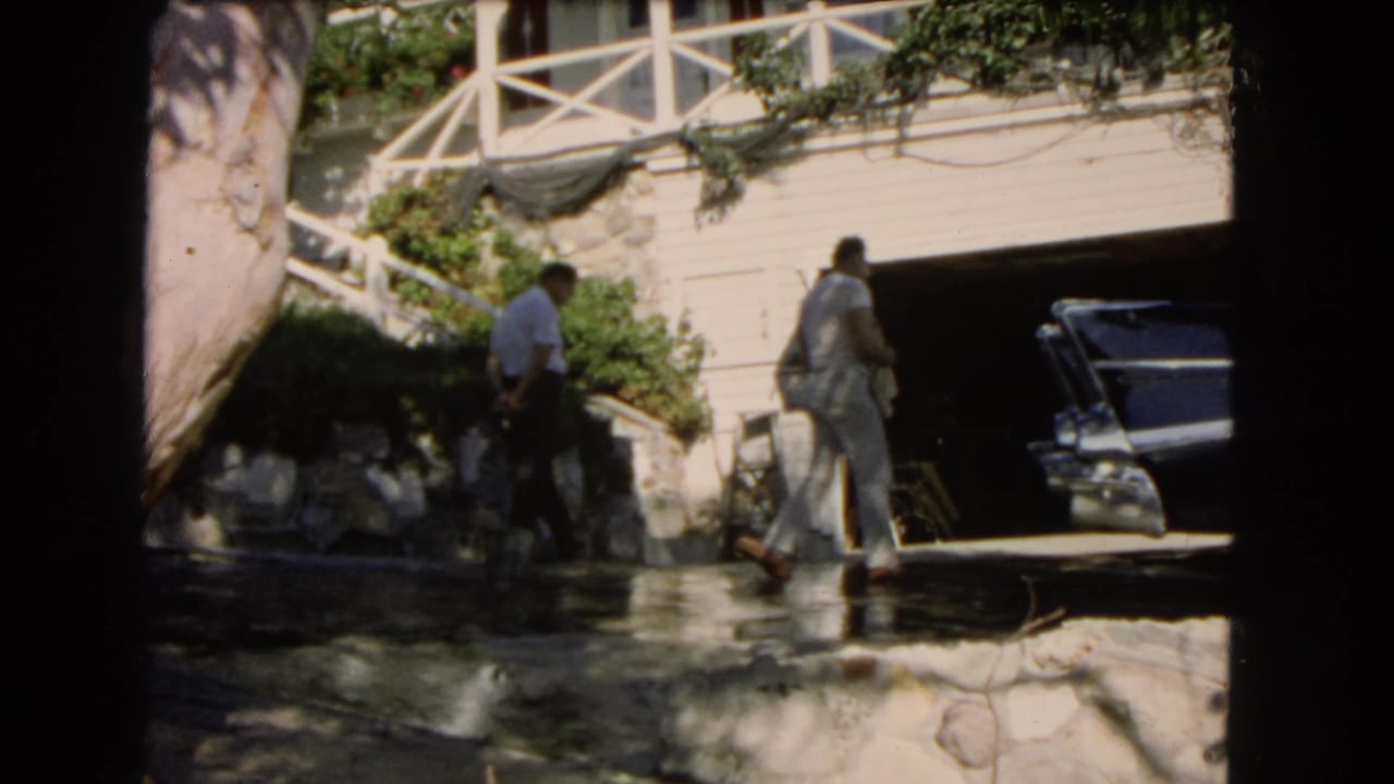 Two men near a driveway, house, stone wall, and dark sedan.