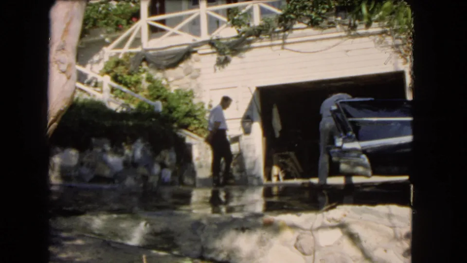 A man stands on the wet driveway while another person works near a dark sedan beside the garage of a hillside Los Angeles home.