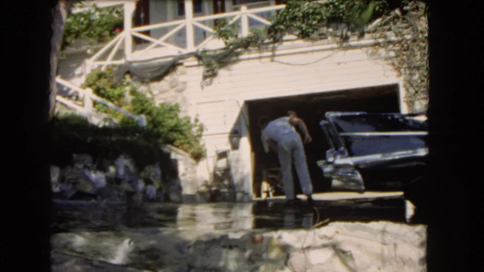 A man bends toward the driveway or garage area beside a dark sedan, with white railings and stone landscaping visible at the residence.