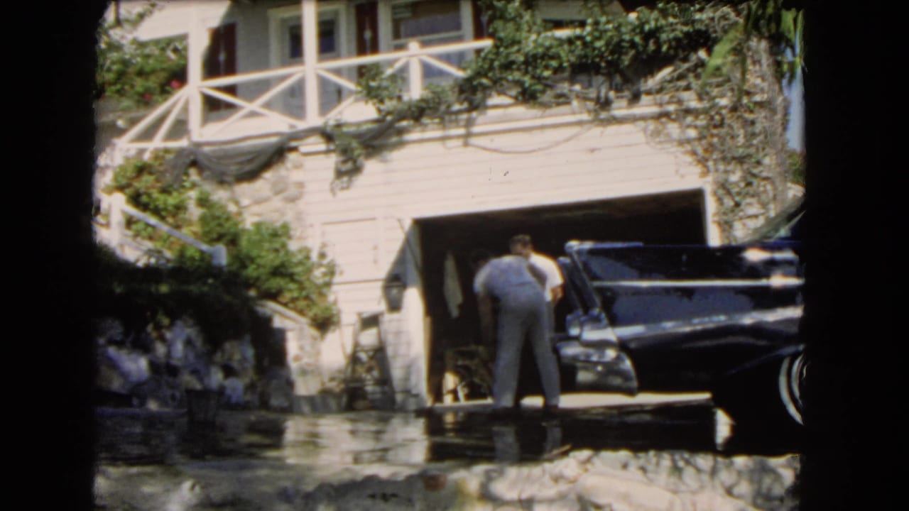 1966 Los Angeles home-movie frame showing two men beside a black classic car near a white garage and sunlit driveway.