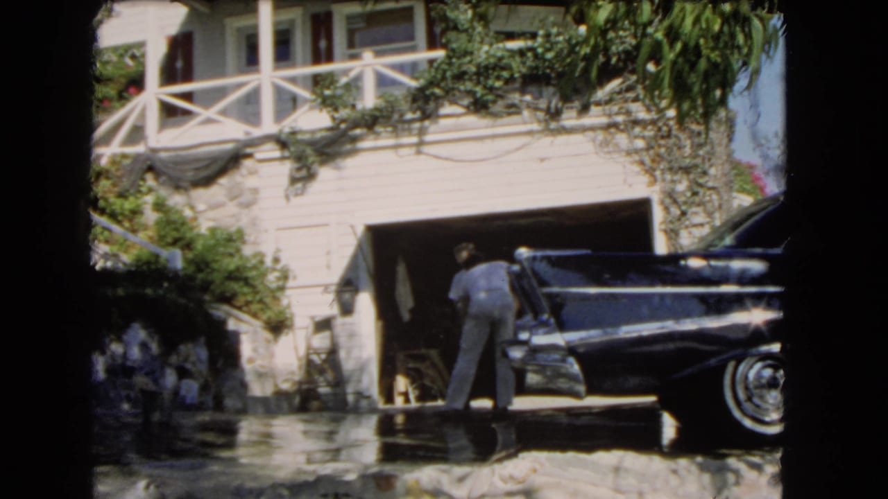A 1966 Los Angeles driveway scene with one man working near a black classic car and another standing beside the garage.