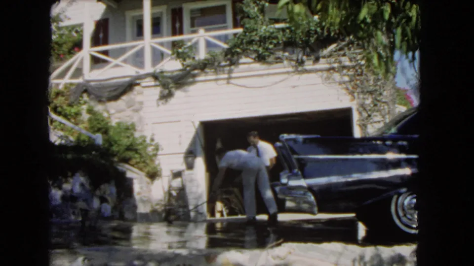 Home-movie frame from 1966 Los Angeles showing two men by a garage and black classic car with greenery across the house.