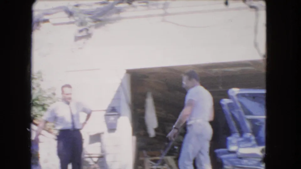 A bright overexposed 1966 Los Angeles home-movie frame shows two men near a garage and the rear section of a dark car.