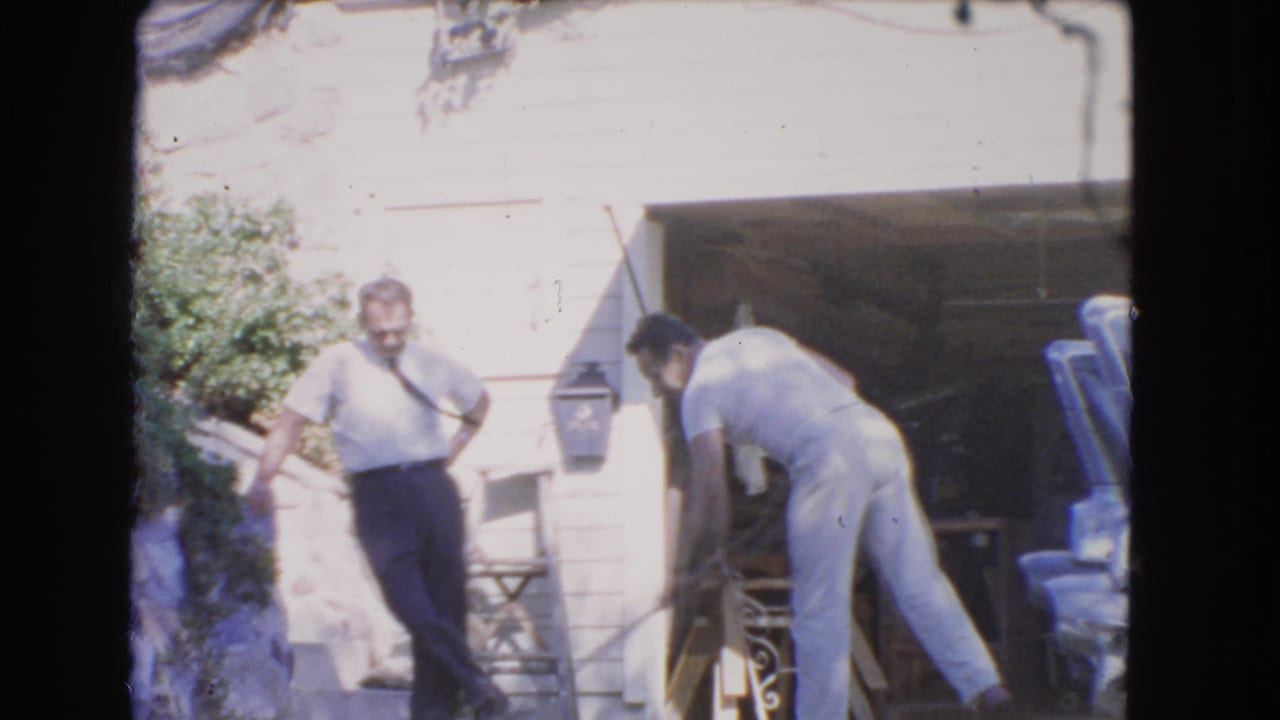 Two adult men outside a house, one standing near a wall while another bends over by a wet driveway.