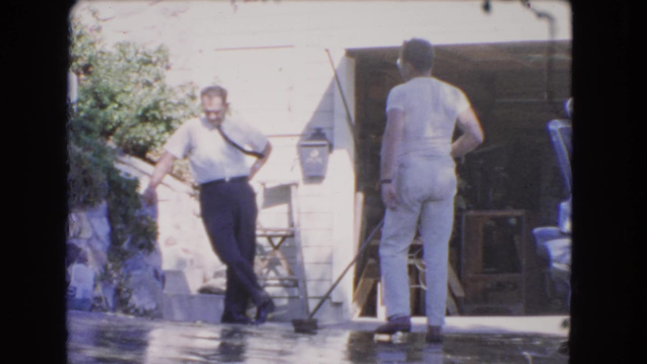 1966 Los Angeles exterior frame with one man standing by the house and another near the open garage, a broom resting on the wet driveway.