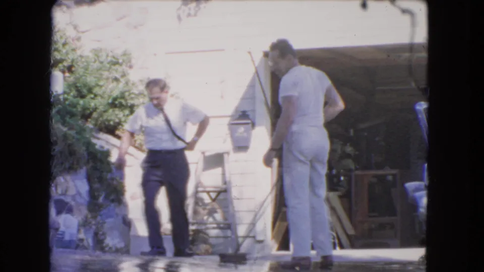 1966 Los Angeles home-movie frame showing two men by a suburban garage, with one walking and the other turned away on the driveway.
