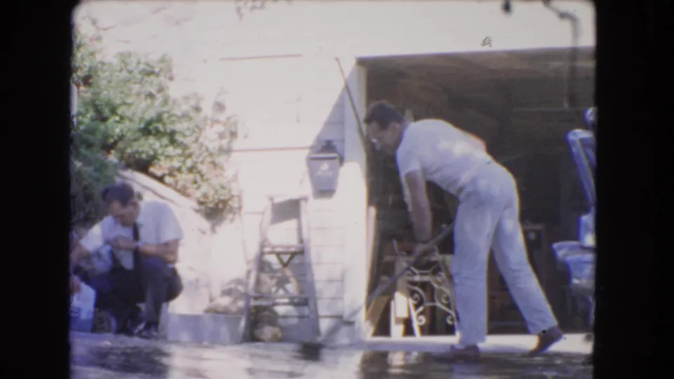 1966 Los Angeles frame showing a man sweeping water on a driveway while another adult man crouches near the house wall.