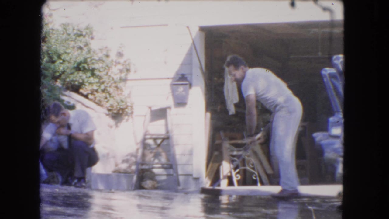 Two men outside a house as one sweeps a wet driveway with a broom.
