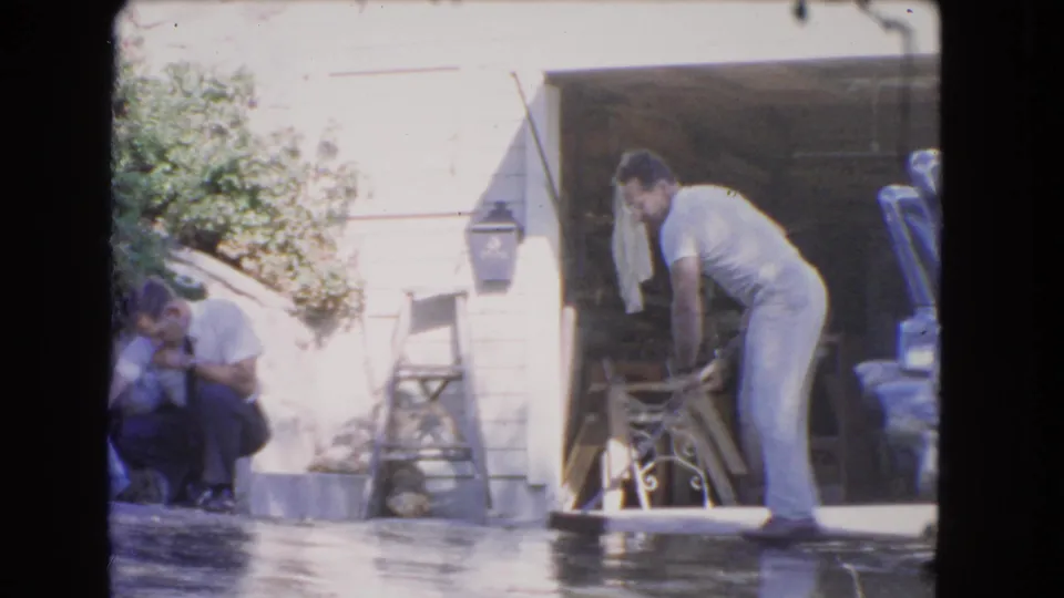 1966 Los Angeles home-movie frame showing one man sweeping a wet driveway while another crouches near the house exterior.