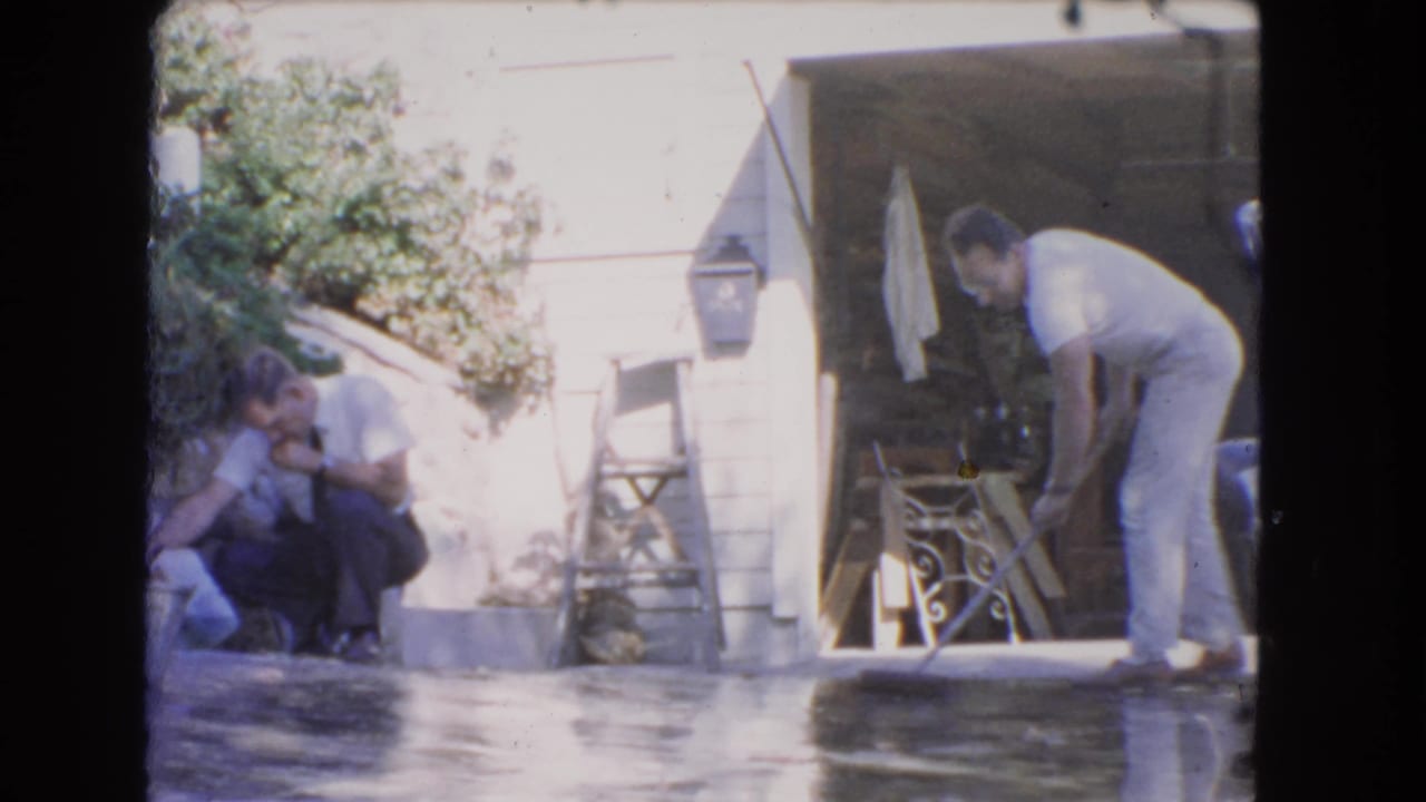 Man sweeping a wet driveway while another man remains near the house.