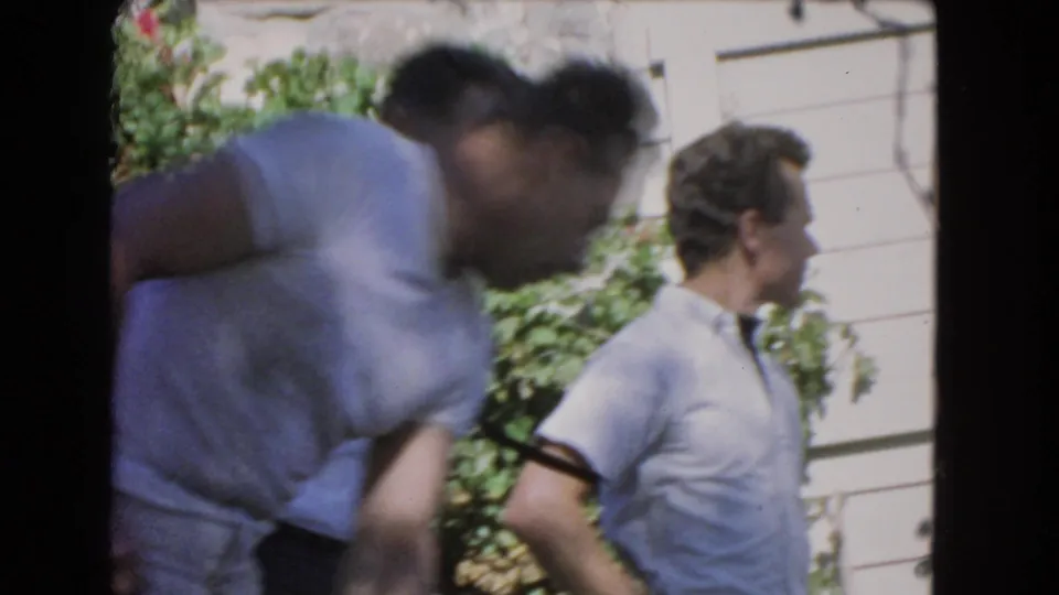 Closer 1966 Los Angeles home-movie frame of two adult men in casual shirts near greenery and the side of a house.