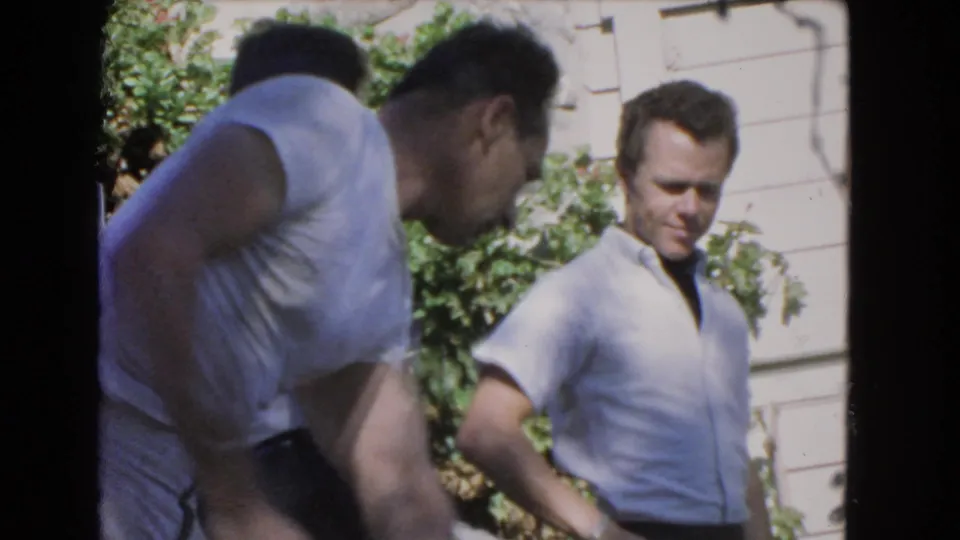 1966 Los Angeles frame showing two adult men near a residential wall and greenery, with one partly blurred in the foreground.