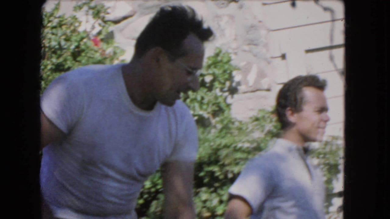 A man in a white T-shirt bends forward while another man stands beside shrubs.